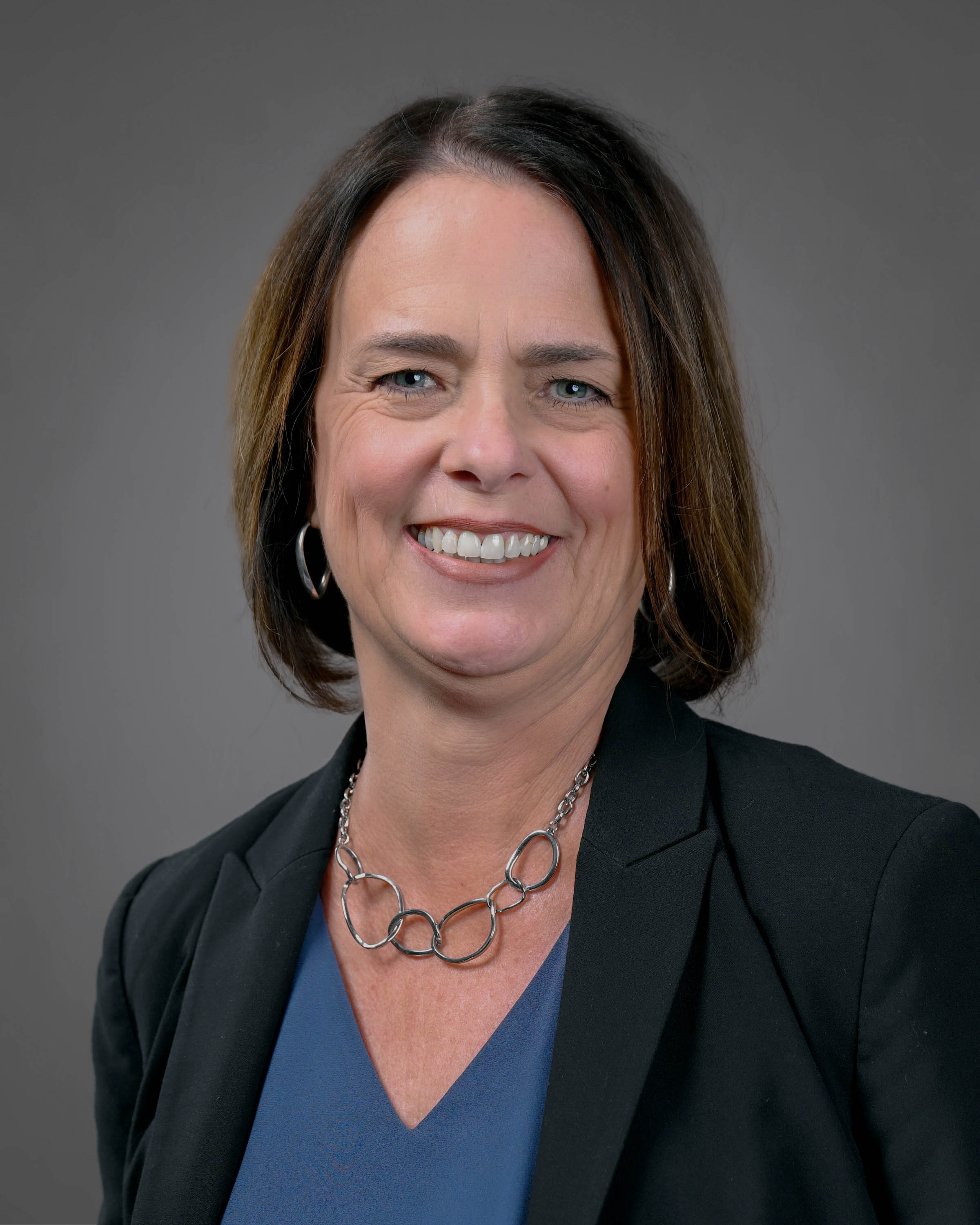 Kind looking woman smiling with brown hair in a black jacket with a dark blue shirt.